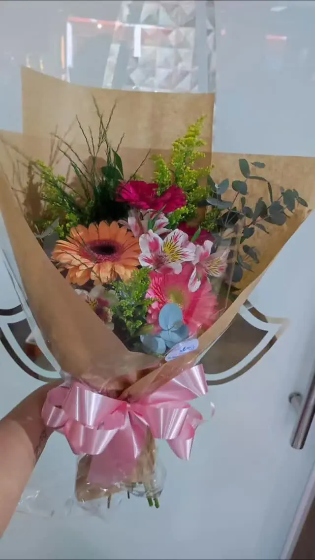 Buquê de gérberas laranja e rosa, astromélias, rosas, tango e eucalipto, embrulhado em papel kraft com laço de cetim rosa, segurado na mão
