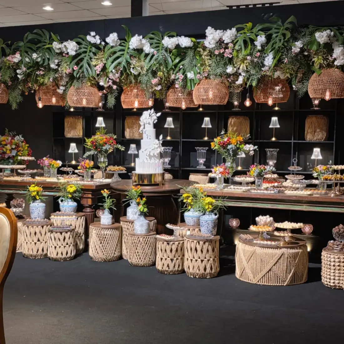 Mesa de doces de casamento sob uma faixa de folhagens com hortênsias brancas e pendentes de palha acesos; bolo branco com as iniciais M e G no centro, doces em bandejas e bancos de corda no chão