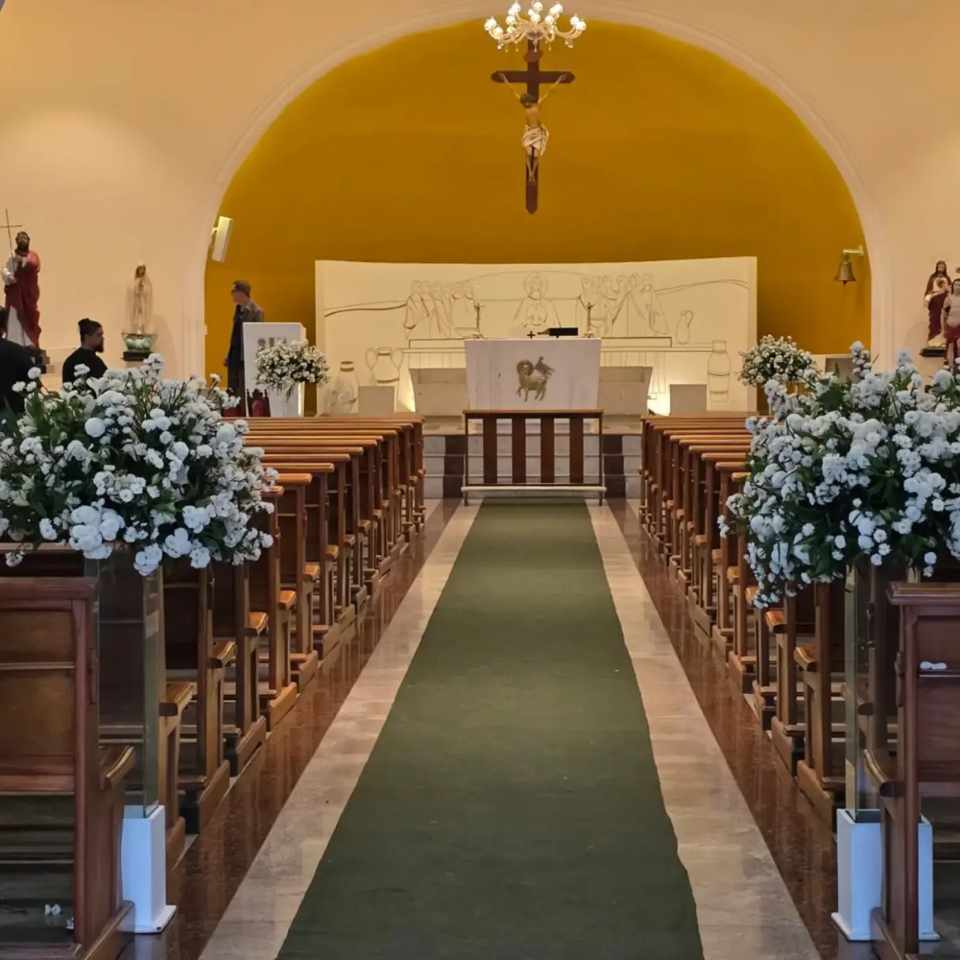 Corredor da Igreja São Filipe decorado para casamento: tapete verde entre os bancos, arranjos de flores brancas em pedestais espelhados ao longo das fileiras e o altar sob o arco amarelo, com o lustre aceso